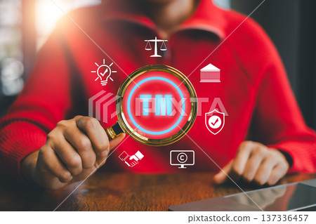 Person in red sweater holding magnifying glass with trademark symbol, surrounded by business icons, emphasizing intellectual property. 137336457