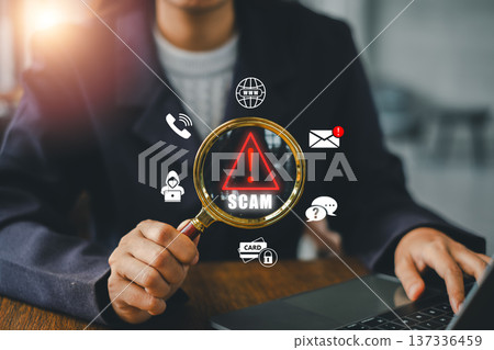 A person examines online scam alerts using a magnifying glass, highlighting digital security icons on a laptop. 137336459