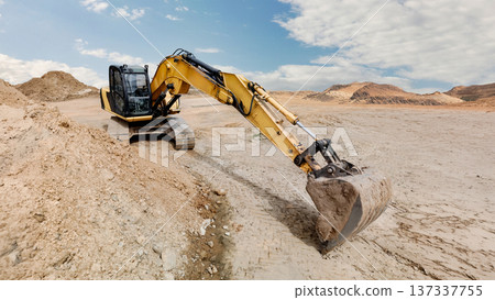A large excavator is digging into dry ground at a construction site. Dust is rising as it moves dirt near hills on a sunny day 137337755