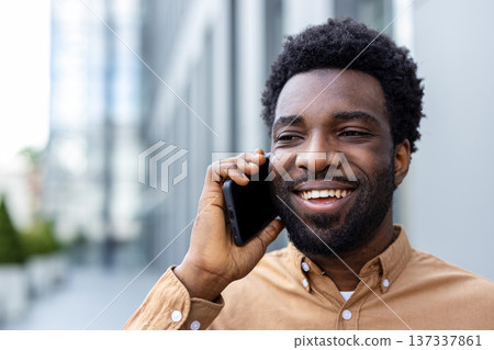Young adult man smiling widely, holding a cellular phone to his ear, enjoying a conversation and communicating while standing in a modern urban environment Young adult man smiling widely, holding a cellular phone to his ear, enjoying a conversation and communicating while standing in a modern urban environment 137337861