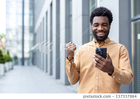 Smiling man feeling excited and making a fist pump gesture, celebrating good news or success received on his mobile phone while standing outdoors in an urban setting 137337864