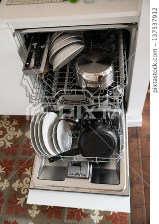Fully loaded dishwasher featuring various dirty kitchen items like plates, bowls, pots, pans, and silverware, representing daily household chores and modern convenience in a kitchen 137337912