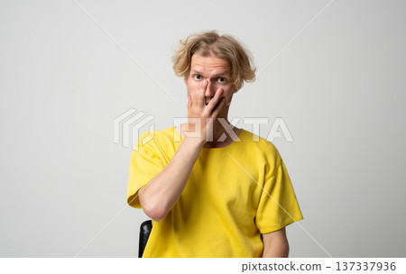 Young blond man expressing shock and disbelief, reacting with wide eyes and covering his face with his hand, conveying surprise, awe, confusion, or a bad outcome 137337936