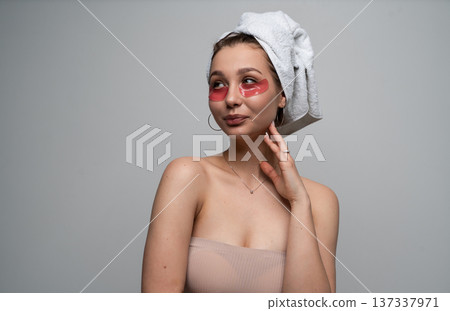 Young woman applying under eye patches and wearing a towel on her head, performing a self-care beauty routine for fresh skin and wellness at home Young woman applying under eye patches and wearing a towel on her head, performing a self-care beauty routine for fresh skin and wellness at home 137337971