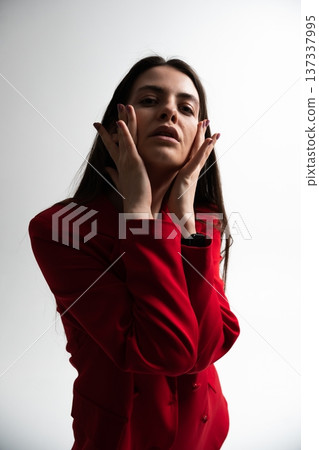 Young adult woman with dark hair touching her face, expressing emotion and beauty in a red fashionable blazer, posing on a studio background with dramatic lighting Young adult woman with dark hair touching her face, expressing emotion and beauty in a red fashionable blazer, posing on a studio background with dramatic lighting 137337995