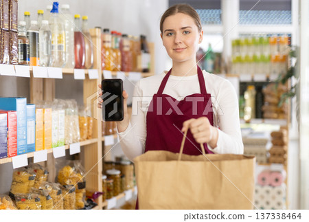 Female seller in uniform with a paper bag showing information on smartphone screen in store 137338464