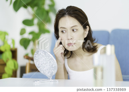 A middle-aged Japanese woman looking in the mirror in the living room and worrying about her skin condition A middle-aged Japanese woman looking in the mirror in the living room and worrying about her skin condition 137338989