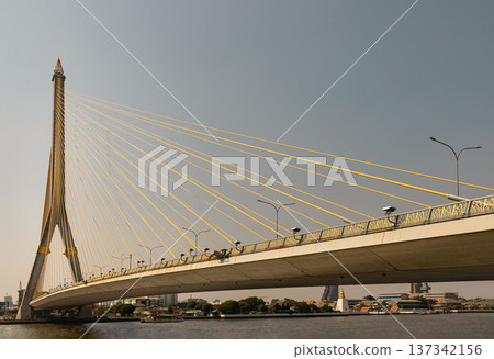 An elegant feat of engineering, the Rama VIII Bridge is anchored by a singular pylon an inverted 'Y' reaching toward the sky with a golden, flame-tipped spire. Golden-yellow cables extend from its An elegant feat of engineering, the Rama VIII Bridge is anchored by a singular pylon an inverted 'Y' reaching toward the sky with a golden, flame-tipped spire. Golden-yellow cables extend from its 137342156