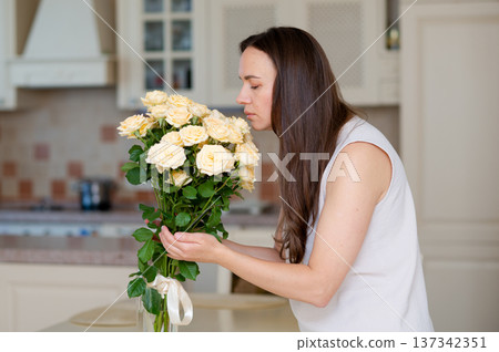 Young woman with long hair smelling beautiful yellow roses in kitchen at home Young woman with long hair smelling beautiful yellow roses in kitchen at home 137342351