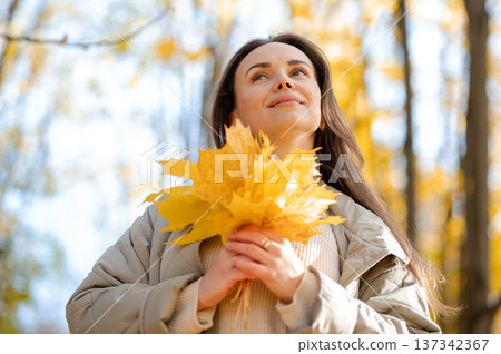 Woman enjoying autumn season holding yellow maple leaves bouquet in sunny fall park Woman enjoying autumn season holding yellow maple leaves bouquet in sunny fall park 137342367