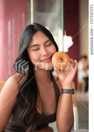 Portrait of a young woman sitting in a cafe with eyes closed holding a glazed donut near her face 137342551