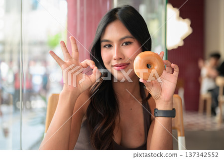 Young Woman Showing OK Gesture While Holding Glazed Donut in Cafe 137342552