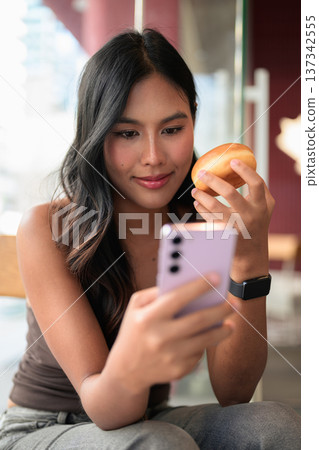 Young Asian Woman Looking at Smartphone While Holding Donut in Cafe Young Asian Woman Looking at Smartphone While Holding Donut in Cafe 137342555