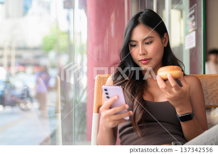 Young Woman Looking at Smartphone While Holding Donut in Cafe Young Woman Looking at Smartphone While Holding Donut in Cafe 137342556