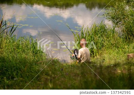 Teenage boy sitting on river bank fishing, relaxing outdoors in summer countryside nature Teenage boy sitting on river bank fishing, relaxing outdoors in summer countryside nature 137342591