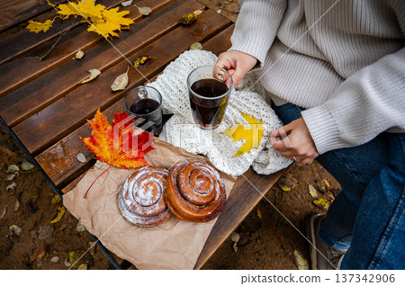 Partial view of woman holding glass cup of coffee beside cinnamon rolls and maple leaves on wooden bench Partial view of woman holding glass cup of coffee beside cinnamon rolls and maple leaves on wooden bench 137342906