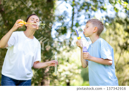 Mother and teen boy having fun blowing soap bubbles in summer park Mother and teen boy having fun blowing soap bubbles in summer park 137342916
