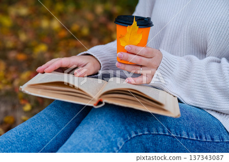 Partial view of woman holding takeaway coffee cup and open book on knees with autumn leaf outdoors in fall park 137343087