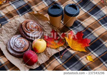 Cozy autumn picnic scene with plaid, two paper cups of hot drinks, pastries, apples and falling maple leave 137343296