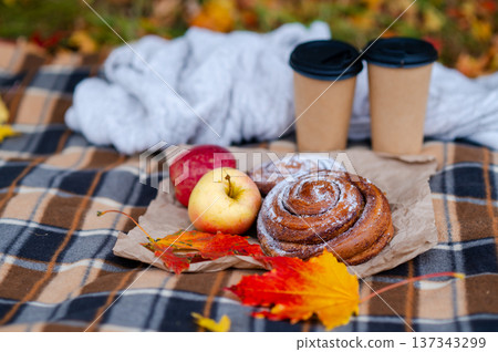 Cozy autumn picnic scene with plaid, knitted sweater, two paper cups of hot drinks, pastries, apples and falling maple leaves 137343299