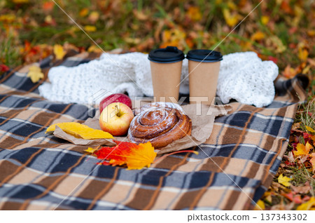 Cozy autumn picnic scene with plaid, knitted sweater, two paper cups of hot drinks, pastries, apples and falling maple leaves 137343302