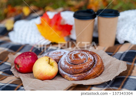Cozy autumn picnic scene with plaid, knitted sweater, two paper cups of hot drinks, pastries, apples and falling maple leaves Cozy autumn picnic scene with plaid, knitted sweater, two paper cups of hot drinks, pastries, apples and falling maple leaves 137343304