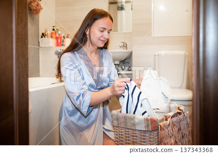 Elegant housewife in silk robe organizing dirty clothes in bathroom, preparing for laundry, concept of home chores and domestic lifestyle 137343368