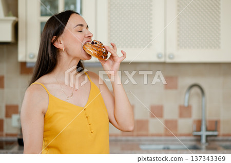 Woman happily biting a sweet bun while standing in the kitchen Woman happily biting a sweet bun while standing in the kitchen 137343369