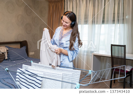 Cheerful housewife listening to music with headphones while hanging laundry on drying rack Cheerful housewife listening to music with headphones while hanging laundry on drying rack 137343410