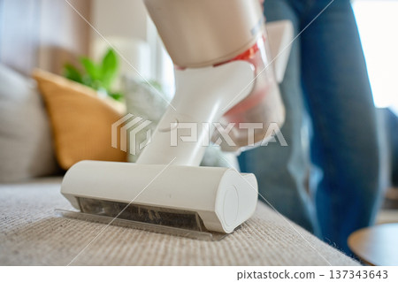 Closeup of handheld vacuum cleaner nozzle on fabric sofa with woman in background Closeup of handheld vacuum cleaner nozzle on fabric sofa with woman in background 137343643