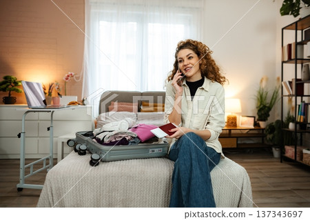 Smiling woman talking on phone while packing suitcase for travel Smiling woman talking on phone while packing suitcase for travel 137343697