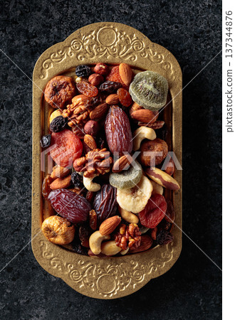 Dried tropical fruits, nuts, and raisins in a golden bowl on black stone table. Dried tropical fruits, nuts, and raisins in a golden bowl on black stone table. 137344876