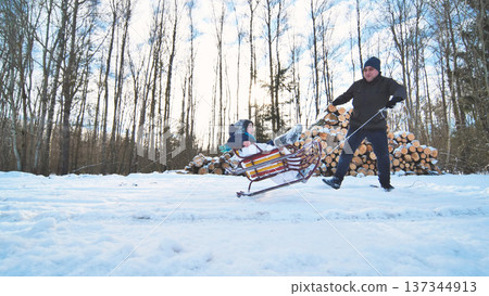 Dad pulls his son on a sled in a snowy forest next to a pile of firewood on a sunny winter day 137344913