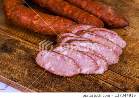 Sliced pieces of cold cuts lie on wooden board with whole sausages nearby in kitchen setting. Sliced pieces of cold cuts lie on wooden board with whole sausages nearby in kitchen setting. 137344919