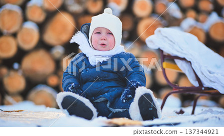Upset toddler wearing warm winter clothes sitting on snow near sled and crying on cold winter day near pile of logs 137344921