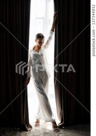 Bride opening curtains in white silk pajamas on wedding day 137344932