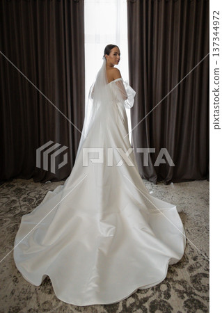 Bride wearing white wedding dress and veil looking out the window in hotel room Bride wearing white wedding dress and veil looking out the window in hotel room 137344972