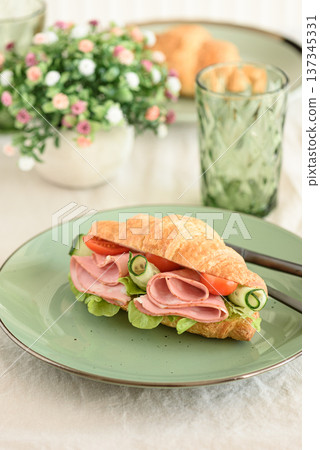 Croissant sandwich filled with ham, lettuce, tomato, and rolled cucumber slices, served on green plate with cutlery Croissant sandwich filled with ham, lettuce, tomato, and rolled cucumber slices, served on green plate with cutlery 137345331