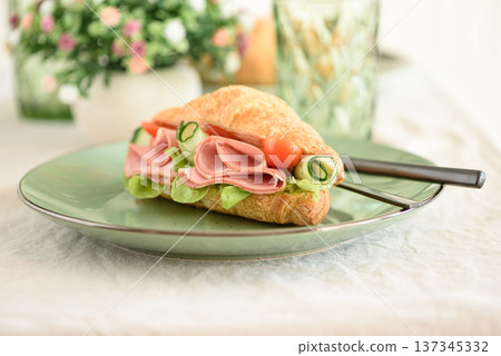 Croissant sandwich filled with ham, lettuce, tomato, and rolled cucumber slices, served on green plate with cutlery Croissant sandwich filled with ham, lettuce, tomato, and rolled cucumber slices, served on green plate with cutlery 137345332
