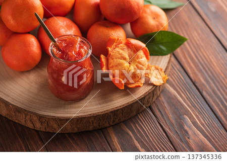 Fresh tangerines with green leaves arranged on  wooden slab, alongside glass jar of homemade jam with golden spoon 137345336