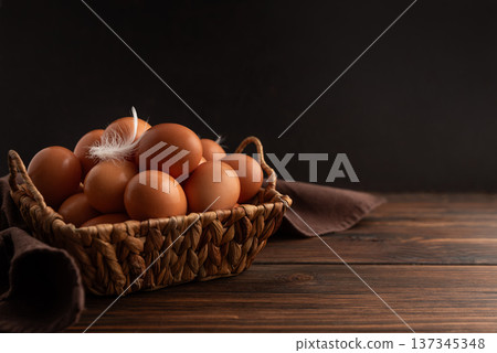 Rustic woven basket filled with fresh brown eggs, placed on wooden background with soft cloth and delicate white feather resting on top 137345348