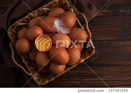 Rustic woven basket filled with fresh brown eggs, placed on wooden background with yalk into shall and delicate white feather resting on top Rustic woven basket filled with fresh brown eggs, placed on wooden background with yalk into shall and delicate white feather resting on top 137345350