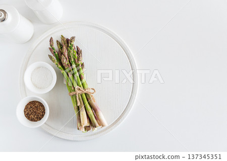 Bundle of fresh asparagus tied with twine on round white wooden tray, accompanied by bowls of coarse salt and ground black pepper 137345351