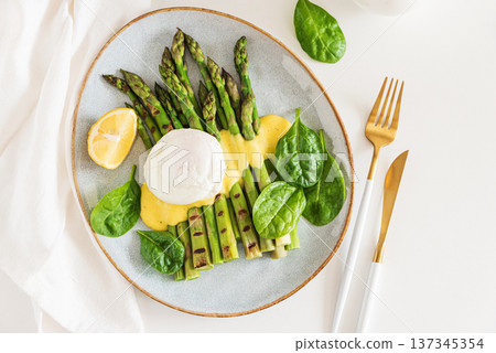 Grilled asparagus spears topped with poached egg and hollandaise sauce, garnished with fresh spinach leaves and lemon wedge 137345354