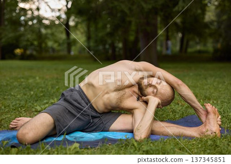 Man without shirt calmly exercises deep breathing on mat in peaceful park Man without shirt calmly exercises deep breathing on mat in peaceful park 137345851