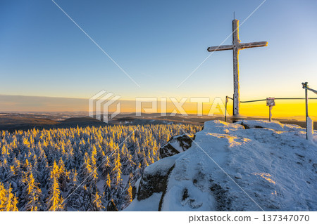 A cross stands on the summit rock of Jizera Mountain. Snow covers the ground and trees. Sunset casts warm light over the landscape, creating a striking contrast with the winter scene. A cross stands on the summit rock of Jizera Mountain. Snow covers the ground and trees. Sunset casts warm light over the landscape, creating a striking contrast with the winter scene. 137347070
