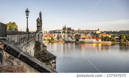 Visitors walk on Charles Bridge while enjoying the view of Prague Castle across the river. The sun shines over the city, creating bright colors in the sky and buildings. Visitors walk on Charles Bridge while enjoying the view of Prague Castle across the river. The sun shines over the city, creating bright colors in the sky and buildings. 137347075