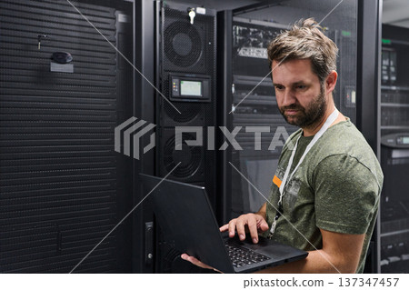 IT technician working on laptop in a data center server room managing network equipment and server racks 137347457
