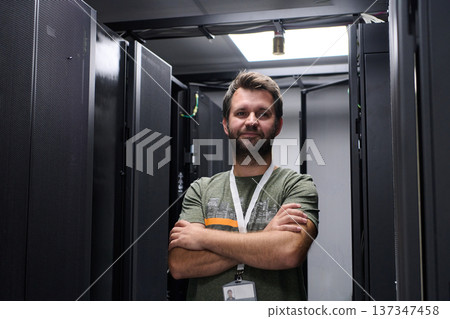 Confident IT technician standing in a data center server aisle with arms crossed and ID badge Confident IT technician standing in a data center server aisle with arms crossed and ID badge 137347458