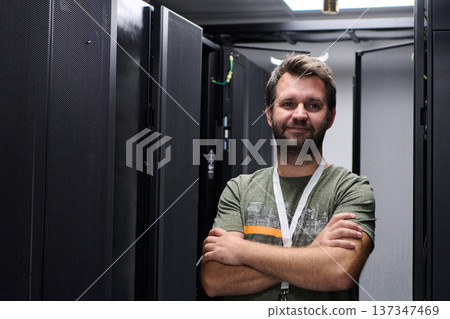 Confident IT technician standing in a data center server room with arms crossed and lanyard 137347469